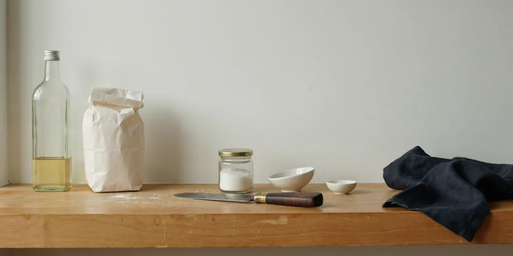 A shelf holding a plain bottle of oil, a paper bag of flour and a jar of salt with completely blank undecorated surfaces, beside one well-made cooking knife with a worn wooden handle