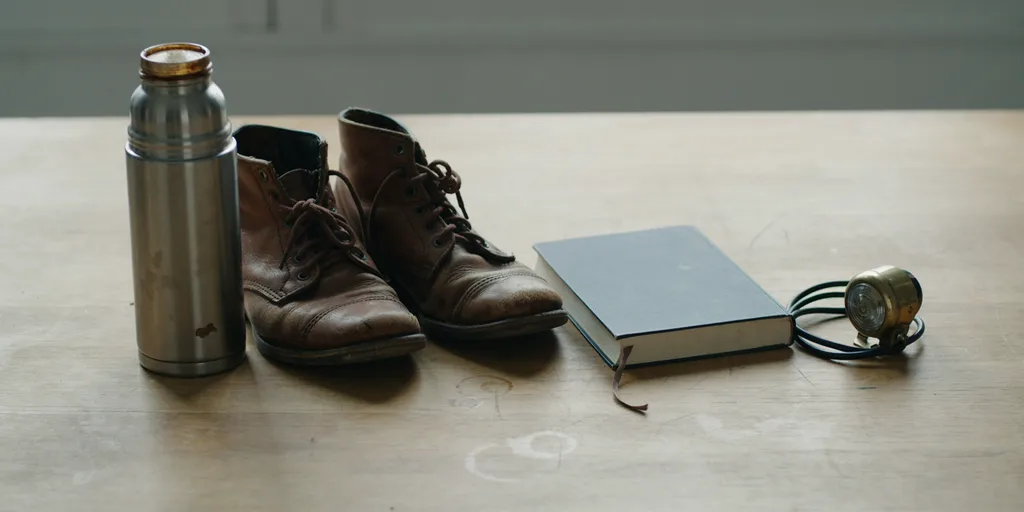 A battered metal thermos flask, a pair of well-worn leather boots, a hardback book with a plain unmarked cover and a small brass bicycle light laid side by side on a worn oak table