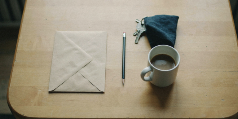 A closed brown envelope, a pencil, a set of keys and a mug of coffee on a worn oak table