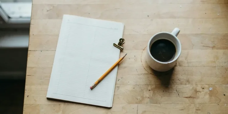 A plain printed planner page with an empty grid on a pale oak table beside a pencil, a brass clip and a mug of coffee
